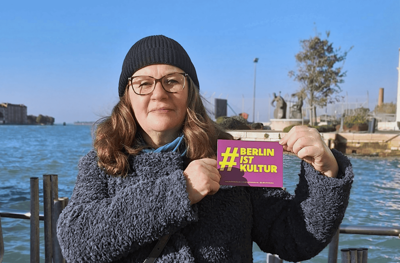 Portrait von Daniela Billig mit einer pinken Postkarte in der Hand, auf der in großen gelben Buchstaben steht: # Berlin Ist Kultur.